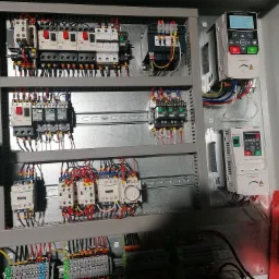 Wnętrze szafy sterowniczej z zainstalowanymi stycznikami Schneider, sterownikami silników Cumark i licznymi przewodami elektrycznymi o różnych kolorach, widoczne oznaczenia i etykiety...