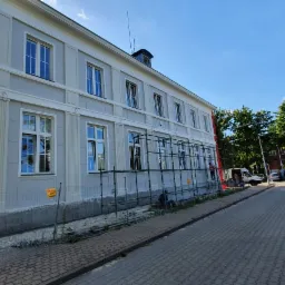 Renowacja elewacji budynku z szarym wykończeniem, widoczne rusztowania i fragment parkingu z samochodami w tle.