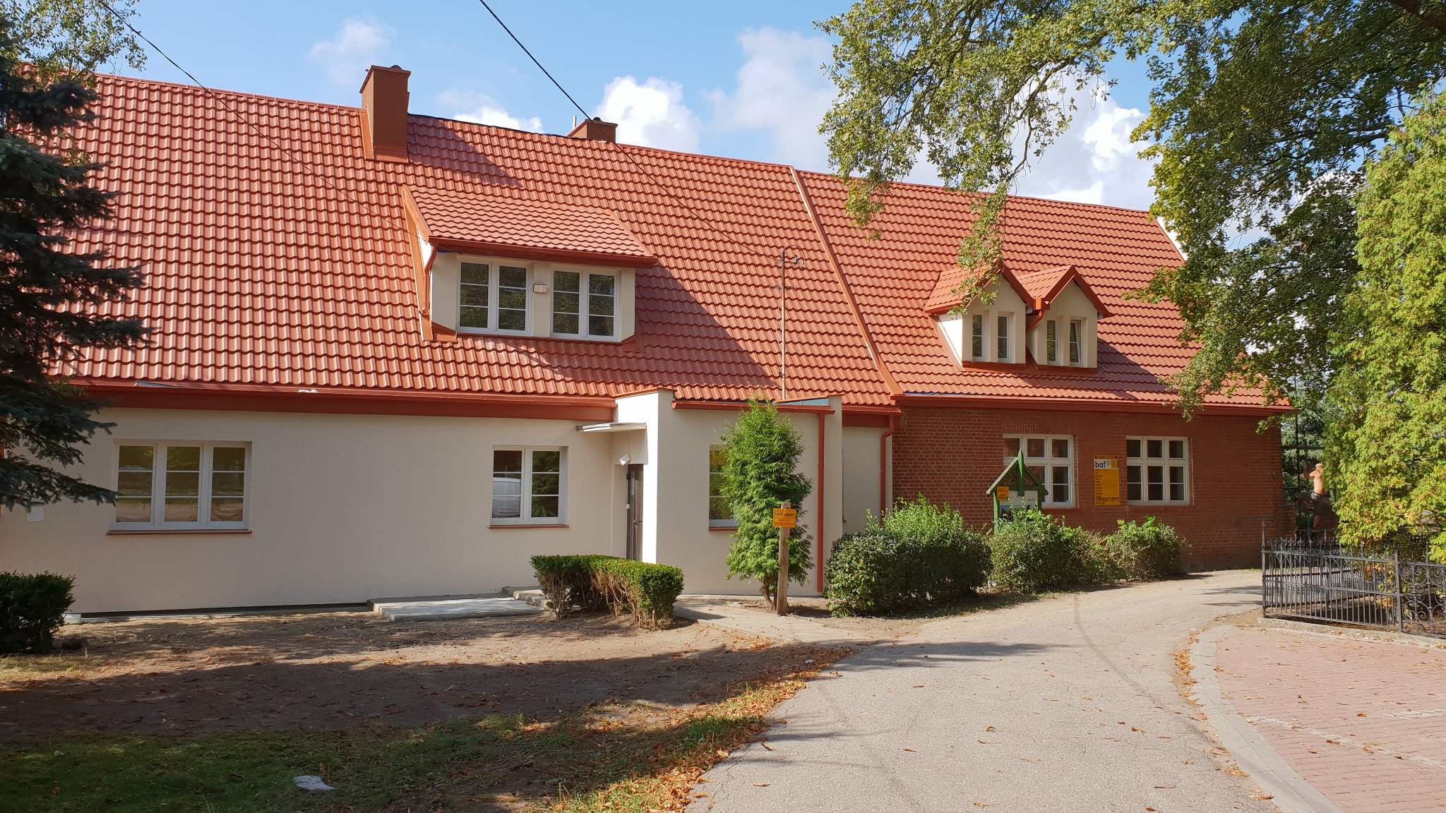 Odnowiony budynek z czerwoną dachówką, kremową elewacją i ceglanym fragmentem, widoczny z poziomu ulicy w słoneczny dzień.
