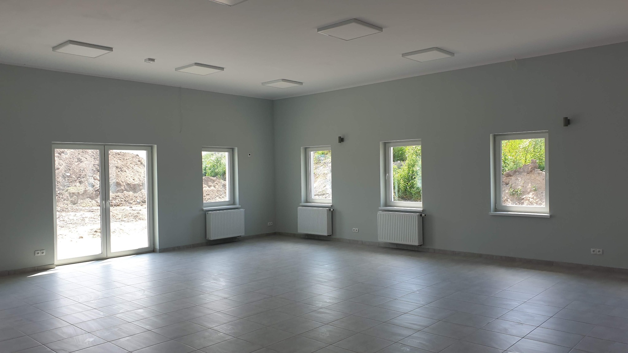 Puste, jasne pomieszczenie z szarymi ścianami, płytkami podłogowymi, białymi grzejnikami pod oknami i kwadratowymi lampami sufitowymi. Widok z okna na ziemię i zieleń.