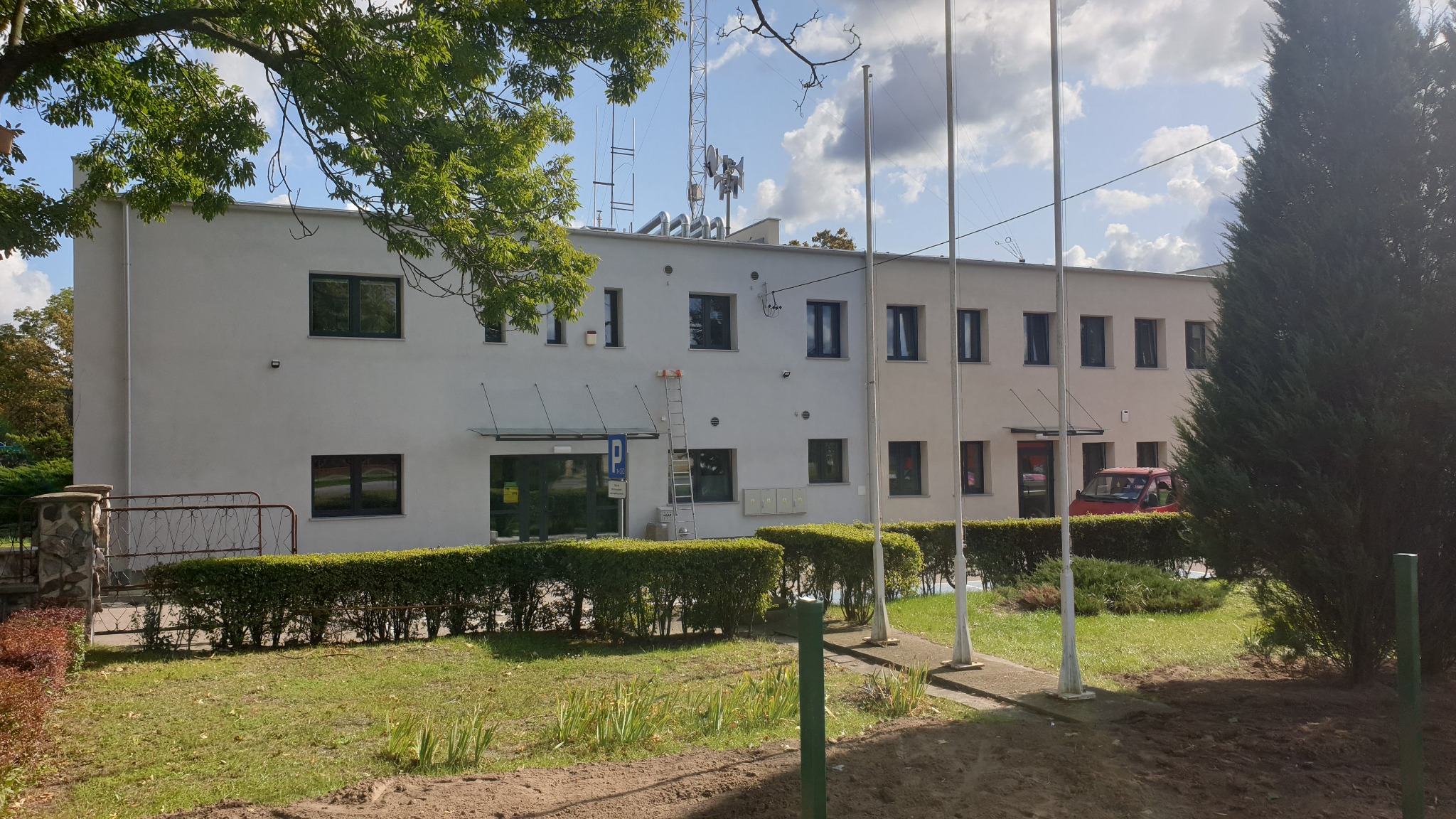 Dwupiętrowy budynek biurowy z jasną elewacją, widoczne okna, wejście z zadaszeniem, antena na dachu i drabina oparta o ścianę, otoczony zielenią.