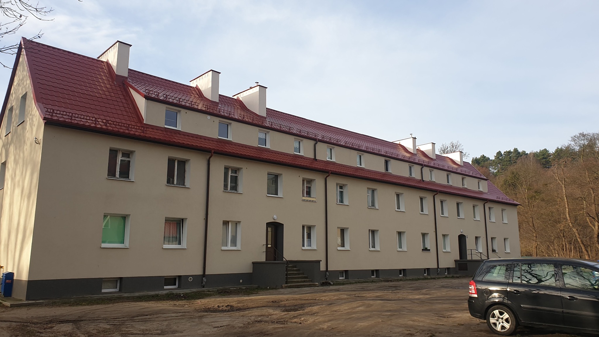 Trzypiętrowy budynek mieszkalny z nową, czerwoną dachówką i beżową elewacją, widok z poziomu gruntu na tle drzew bez liści i fragmentu czarnego samochodu.