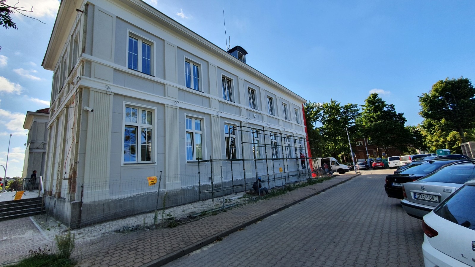 Renowacja elewacji budynku z szarym wykończeniem, widoczne rusztowania i fragment parkingu z samochodami w tle.