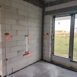 Wnętrze w stanie surowym: instalacja elektryczna w ścianach z betonu komórkowego z widocznymi puszkami instalacyjnymi, przewodami oraz peszlami podłogowymi, okno z widokiem na zewnątrz.