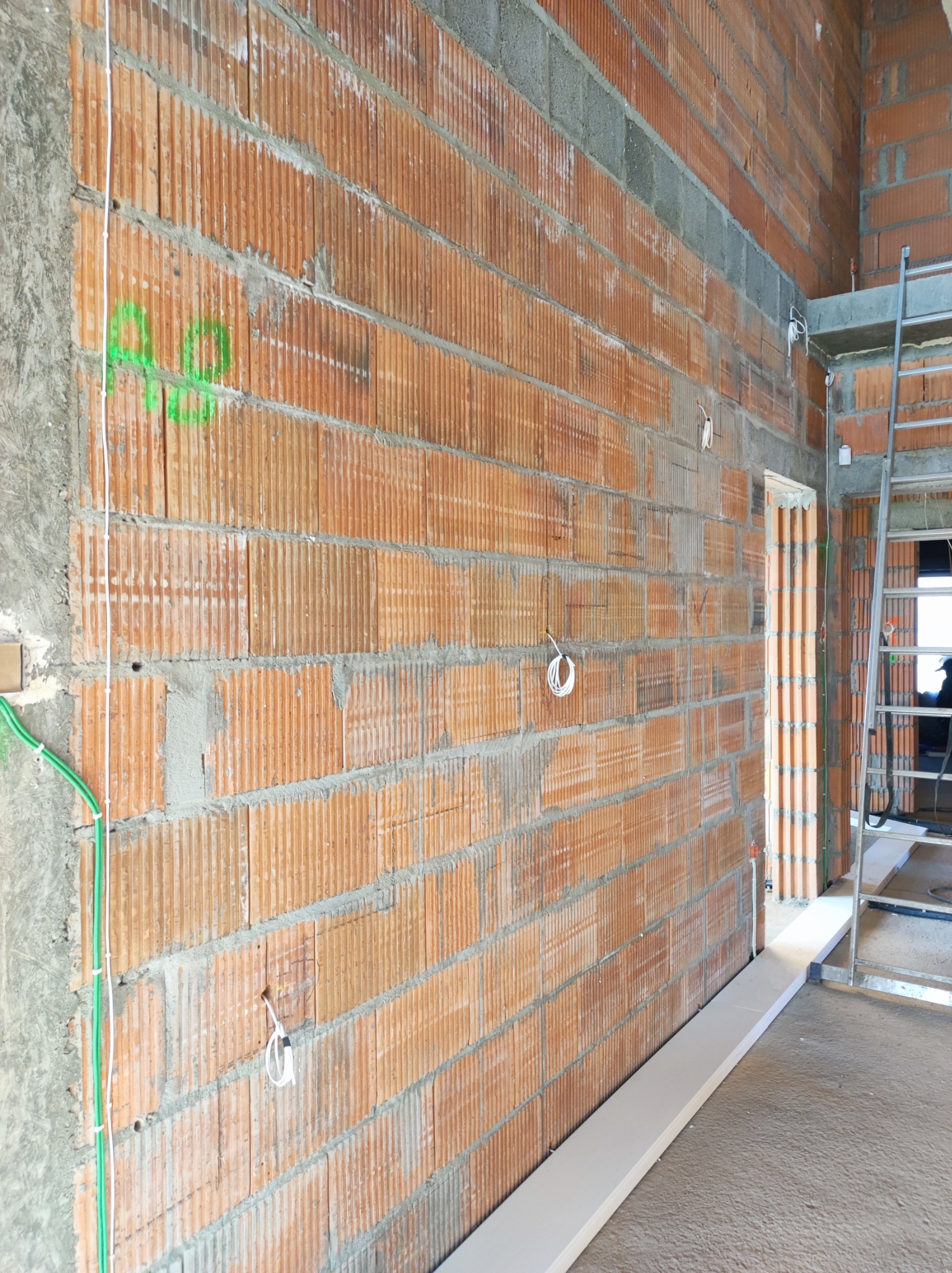 Czerwona ściana z pustaków ceramicznych z widocznymi przewodami elektrycznymi wystającymi ze ściany, zielone oznaczenia na murze, drabina aluminiowa w tle.