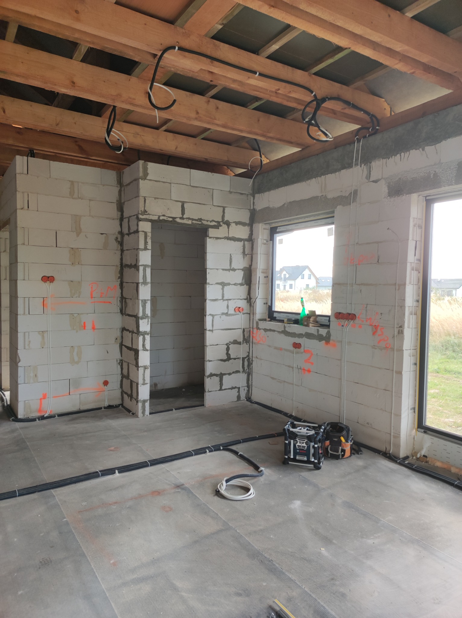 Wnętrze surowego domu w trakcie prac elektrycznych: widoczne przewody rozprowadzone po suficie i ścianach z betonu komórkowego, oznaczenia tras kabli pomarańczowym sprayem, skrzynka narzędziowa...