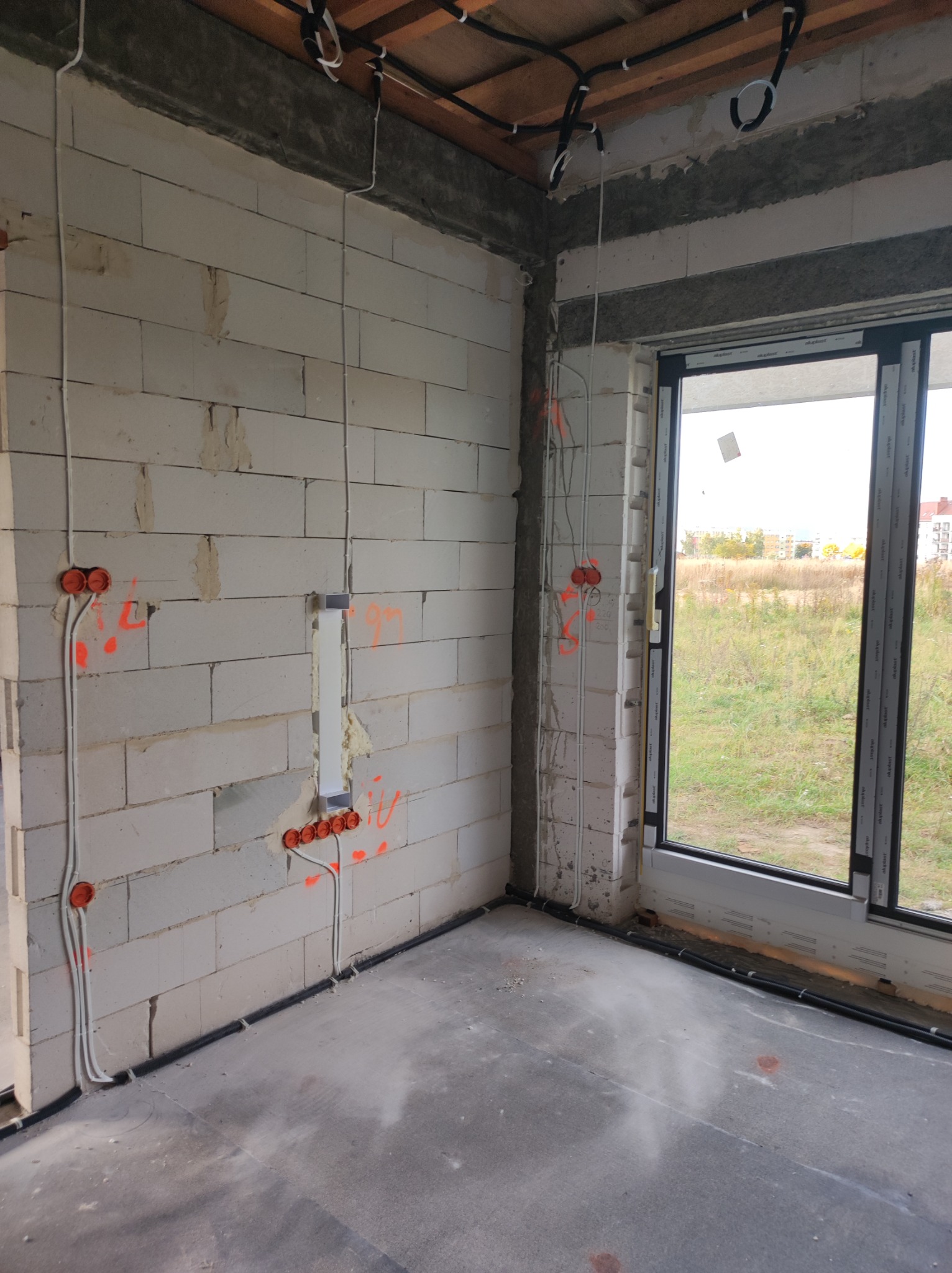 Wnętrze w stanie surowym: instalacja elektryczna w ścianach z betonu komórkowego z widocznymi puszkami instalacyjnymi, przewodami oraz peszlami podłogowymi, okno z widokiem na zewnątrz.