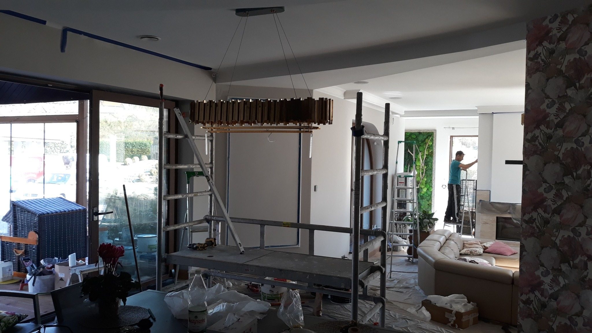 Wnętrze w trakcie remontu: widoczna drabina, rusztowanie, nowoczesna lampa wisząca, malarz na drabinie malujący ścianę, zabezpieczona folią podłoga, oklejone taśmą malarską ściany.