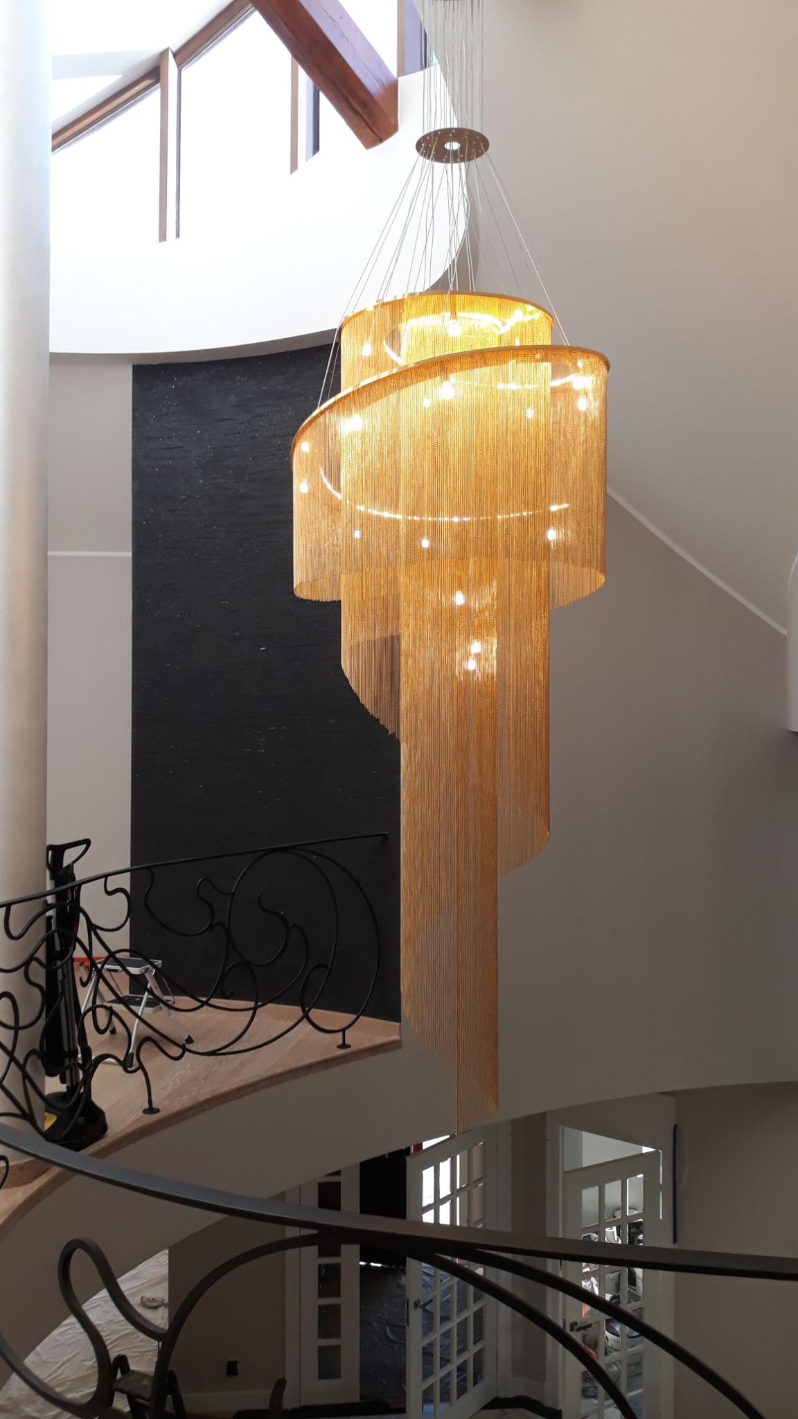 Trzykondygnacyjny, złoty żyrandol kaskadowy z łańcuszków, zainstalowany w przestronnym holu z kolumną i spiralnymi schodami z kutą balustradą.