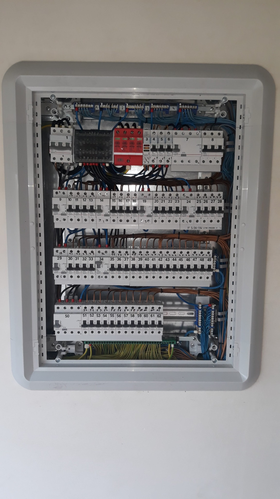 Rozdzielnica elektryczna w obudowie, widoczne zabezpieczenia nadprądowe, różnicowoprądowe, przewody w kolorach niebieskim, brązowym i żółto-zielonym, numery identyfikacyjne na wyłącznikach