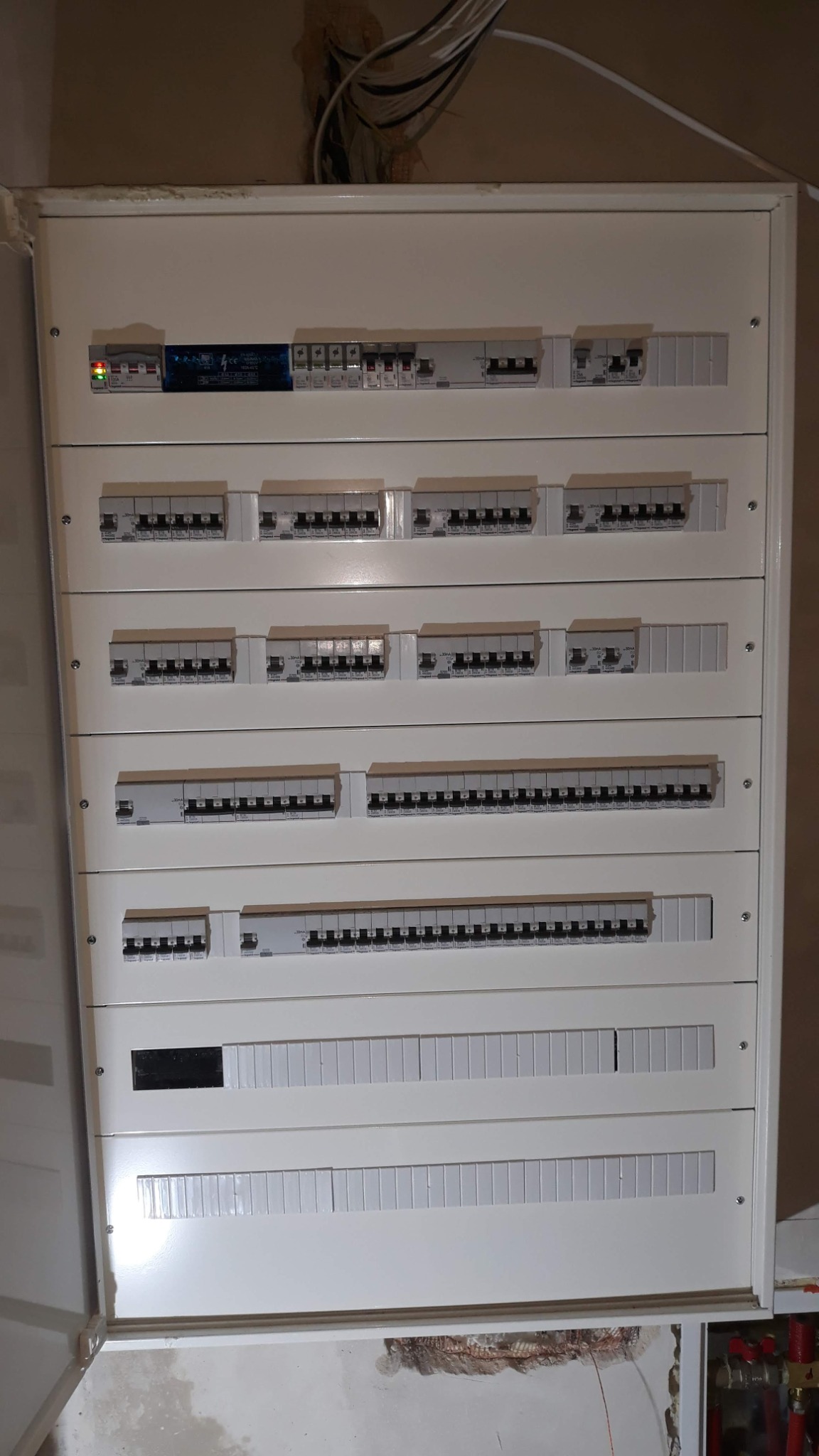 Rozdzielnica elektryczna z wieloma rzędami zabezpieczeń różnicowoprądowych, zamontowana na ścianie w trakcie prac wykończeniowych, widoczne kable wystające ze ściany powyżej.