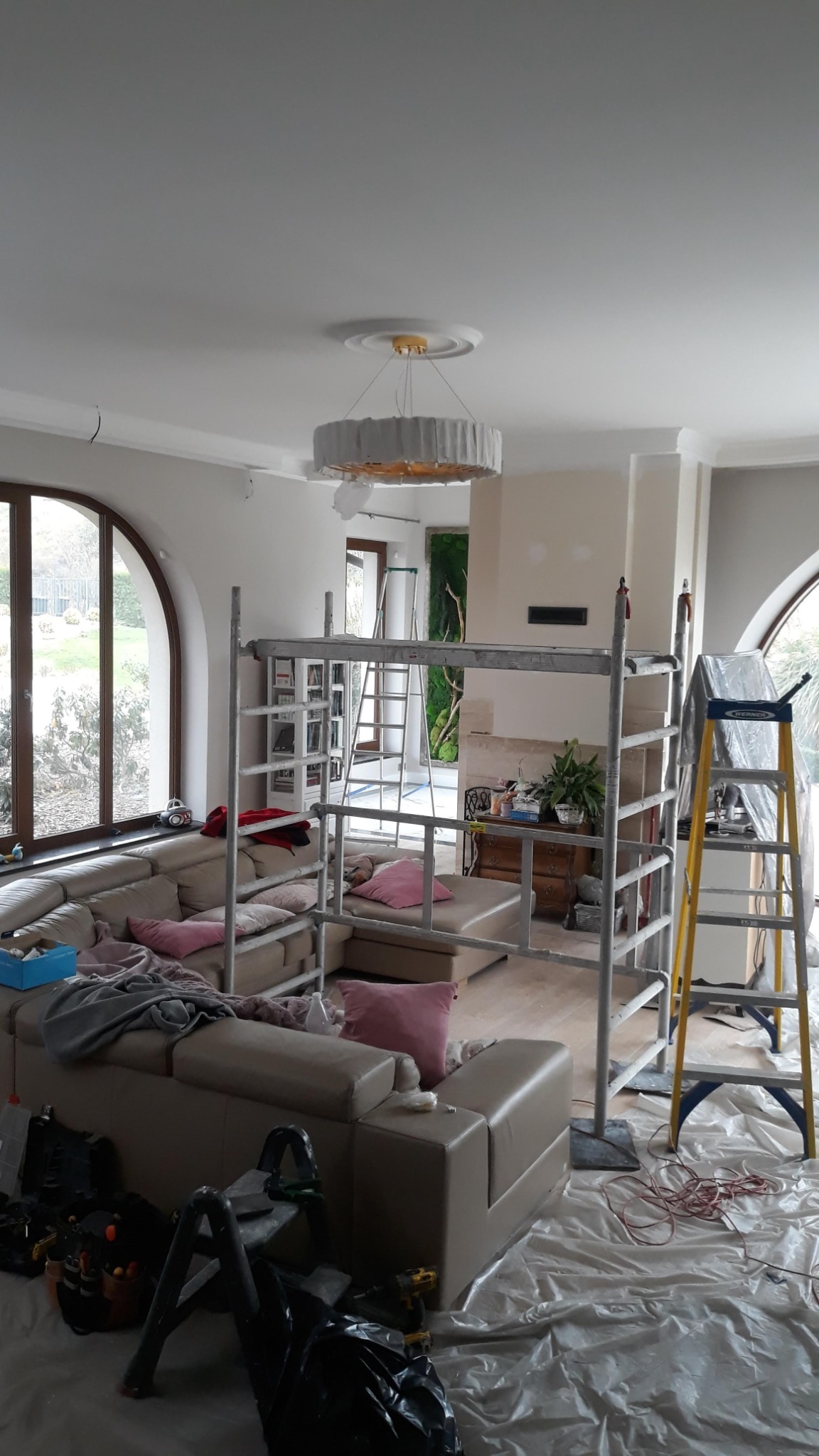 Montaż okrągłej lampy sufitowej z białym abażurem w salonie, widoczne rusztowanie, drabina i zabezpieczona folią podłoga.