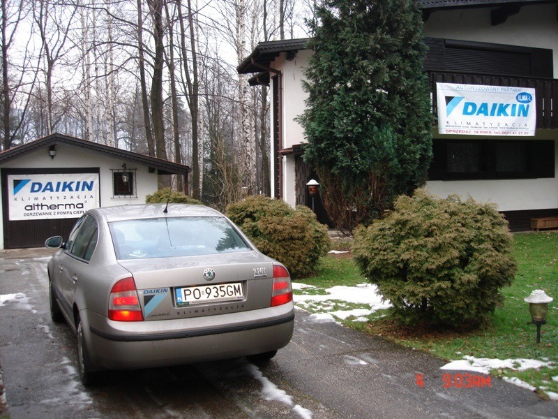 Srebrny samochód Skoda Octavia z naklejką firmy klimatyzacyjnej zaparkowany na podjeździe przed budynkiem z banerami reklamowymi tej samej firmy, widoczne logo Daikin.