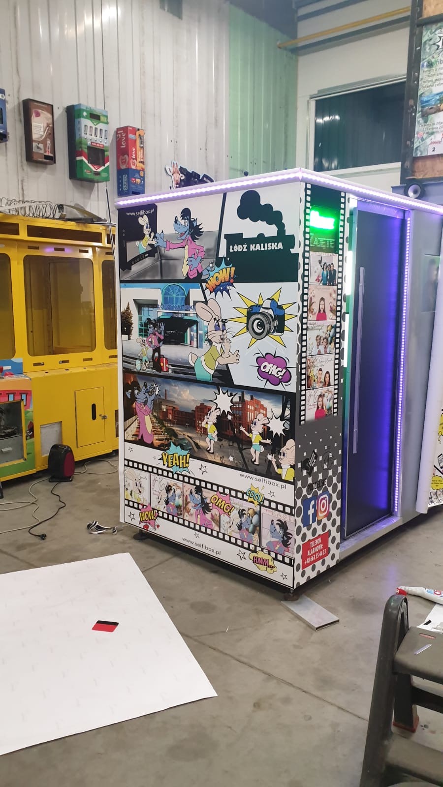 Oklejona fotobudka z motywami komiksowymi i zdjęciami, z adresem strony internetowej i numerem telefonu firmy z Świdnicy, znajdująca się w hali produkcyjnej.