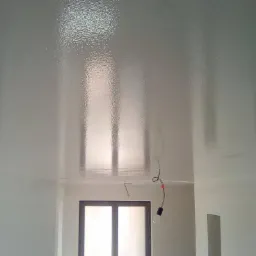 Biały, błyszczący sufit po gładzi, z widocznymi odbiciami światła i elementami instalacji elektrycznej. Widoczne okno i fragment rusztowania.