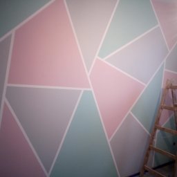 Mirmark Marek Kopeć - Ściana z geometrycznym wzorem w pastelowych kolorach, oddzielona białymi liniami. W rogu drabina malarska. Efekt malowania dekoracyjnego.