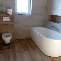 Wykończona łazienka z wanną, toaletą i drewnopodobnymi płytkami na podłodze i ścianie. Widoczne detale: armatura, okno z akcesoriami, półka w ścianie.