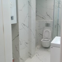 Mirmark Marek Kopeć - Wykończona łazienka w bieli z marmurowym wzorem na płytkach, widoczna kabina prysznicowa, toaleta i umywalka. Na ścianie piec gazowy. Całość w nowoczesnym stylu.