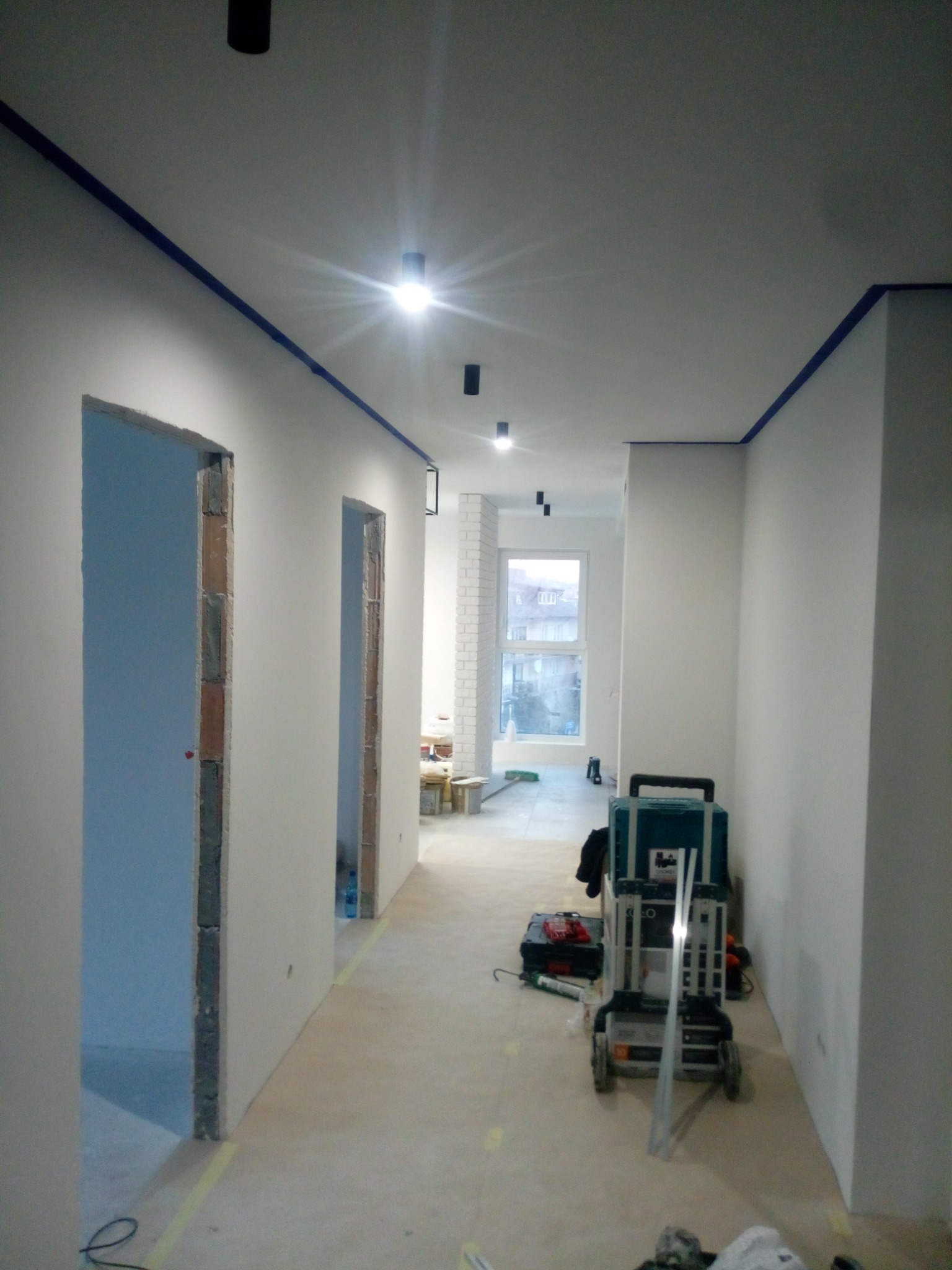 Korytarz w trakcie remontu: ściany po szpachlowaniu i malowaniu na biało, z widocznymi otworami drzwiowymi, zabezpieczonymi taśmą sufitami, narzędziami i materiałami budowlanymi na podłodze.