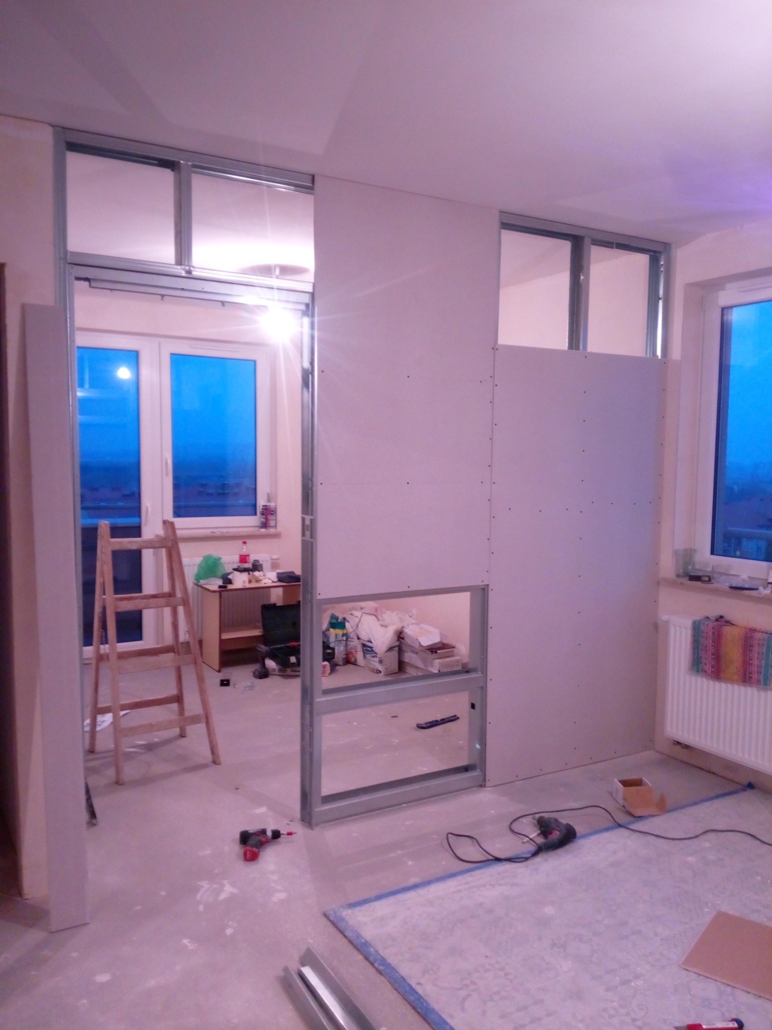 Montaż ścianki działowej z płyty gipsowo-kartonowej na metalowym stelażu, widoczne narzędzia i drabina w tle, okno z widokiem na miasto.