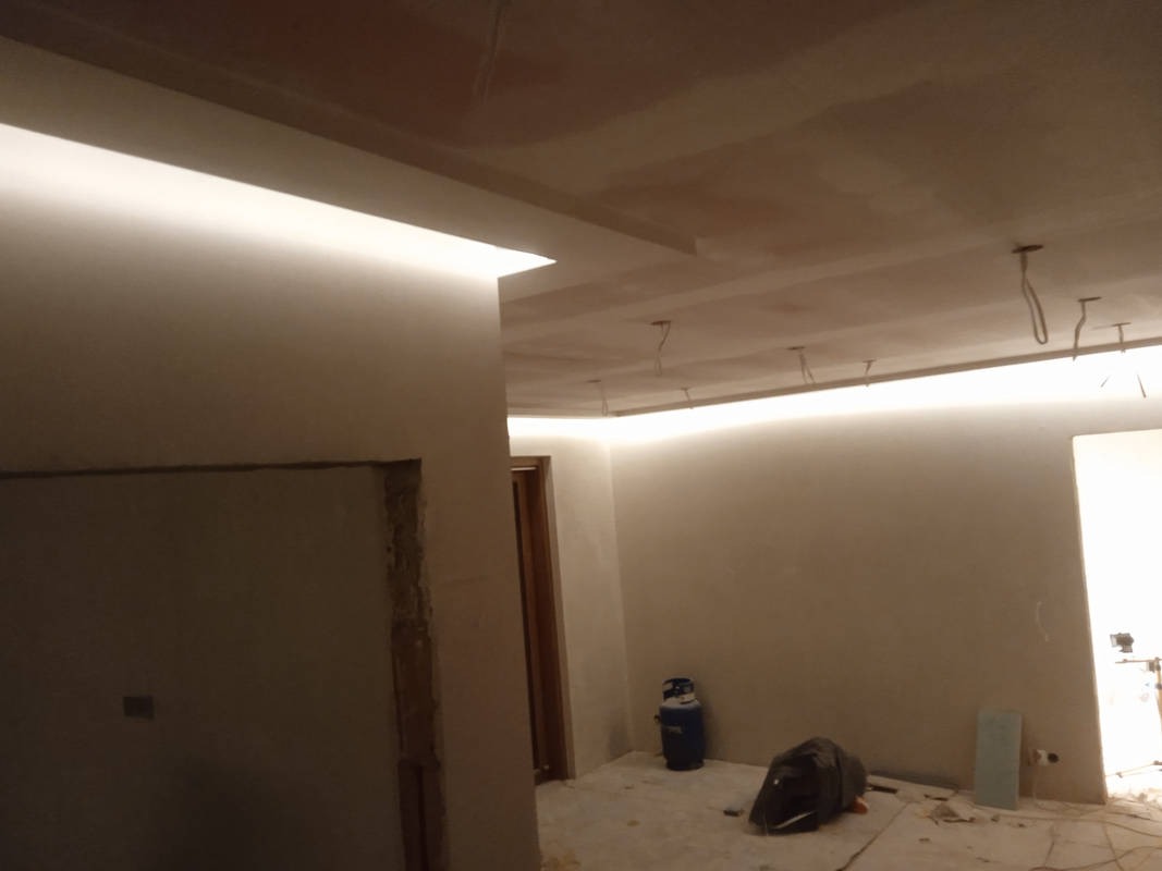 Wnętrze w trakcie remontu z oświetleniem LED na suficie podwieszanym. Widoczne niedoskonałości ścian i sufitu przed wykończeniem. Gazowa butla i worki budowlane w tle.