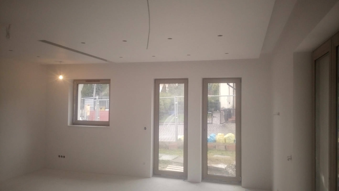 Jasne wnętrze po remoncie: białe ściany, sufit z podwieszanymi elementami, okna i drzwi balkonowe z widokiem na ogród. Minimalistyczny design, świeżo po pracach wykończeniowych.