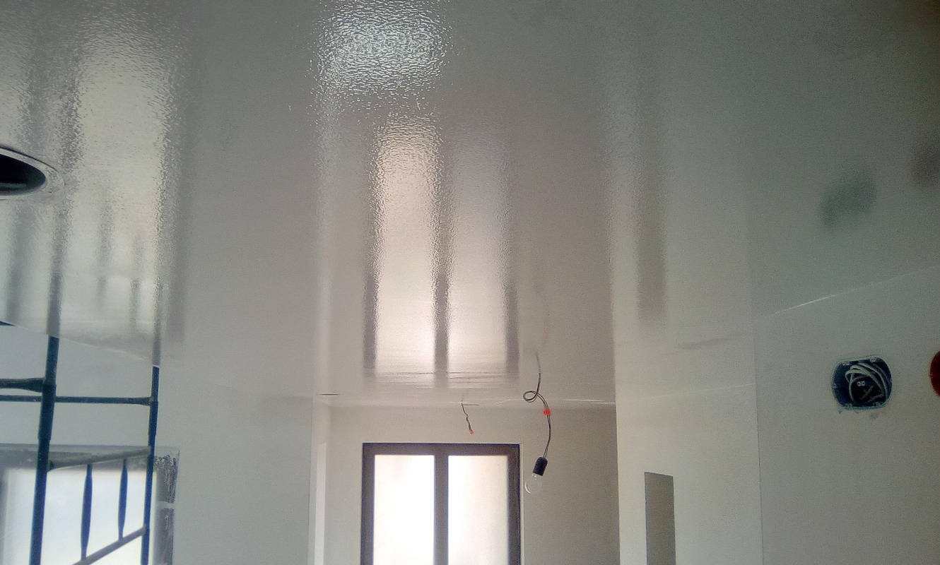 Biały, błyszczący sufit po gładzi, z widocznymi odbiciami światła i elementami instalacji elektrycznej. Widoczne okno i fragment rusztowania.