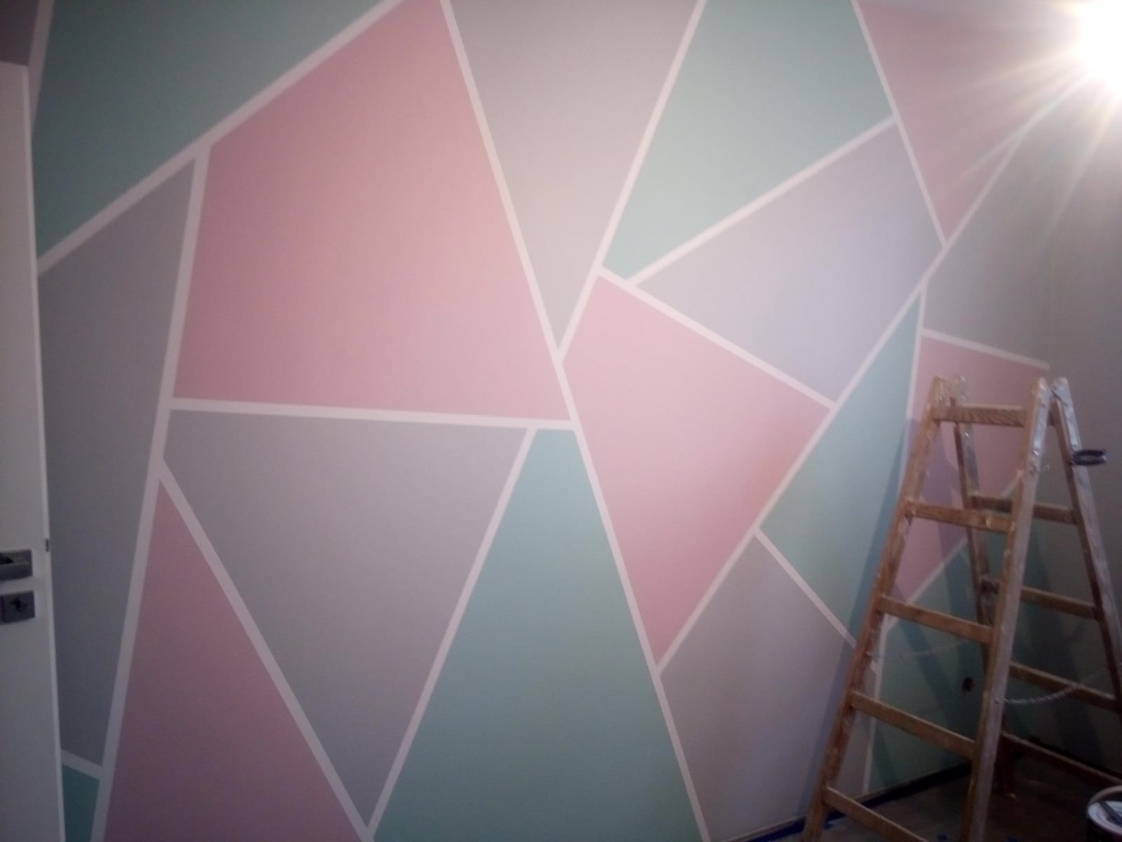 Ściana z geometrycznym wzorem w pastelowych kolorach, oddzielona białymi liniami. W rogu drabina malarska. Efekt malowania dekoracyjnego.