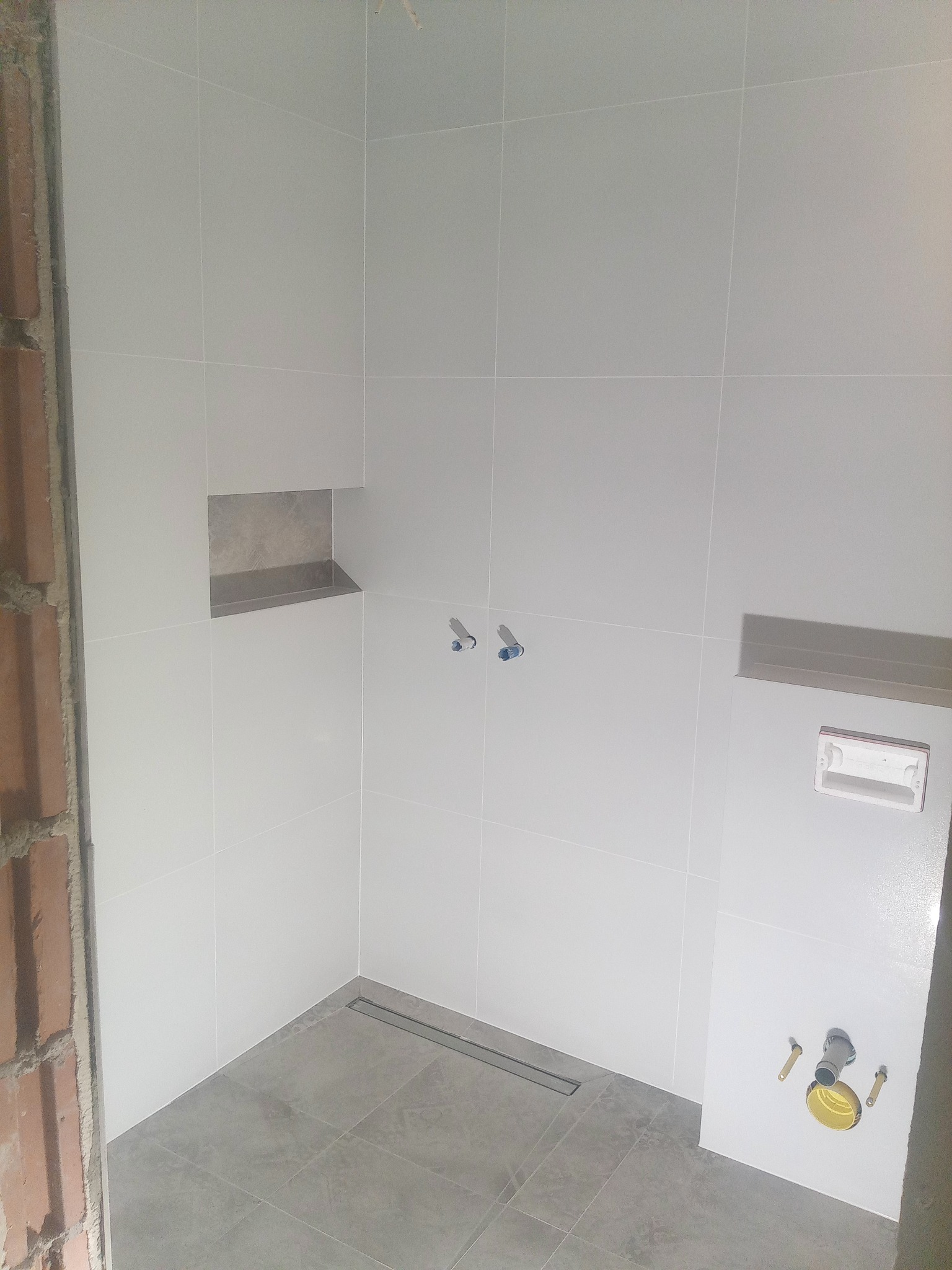 Wnętrze łazienki w trakcie wykańczania: szare płytki podłogowe i białe ścienne, widoczne elementy instalacji wodnej i odpływ liniowy w podłodze. Surowa cegła obok.