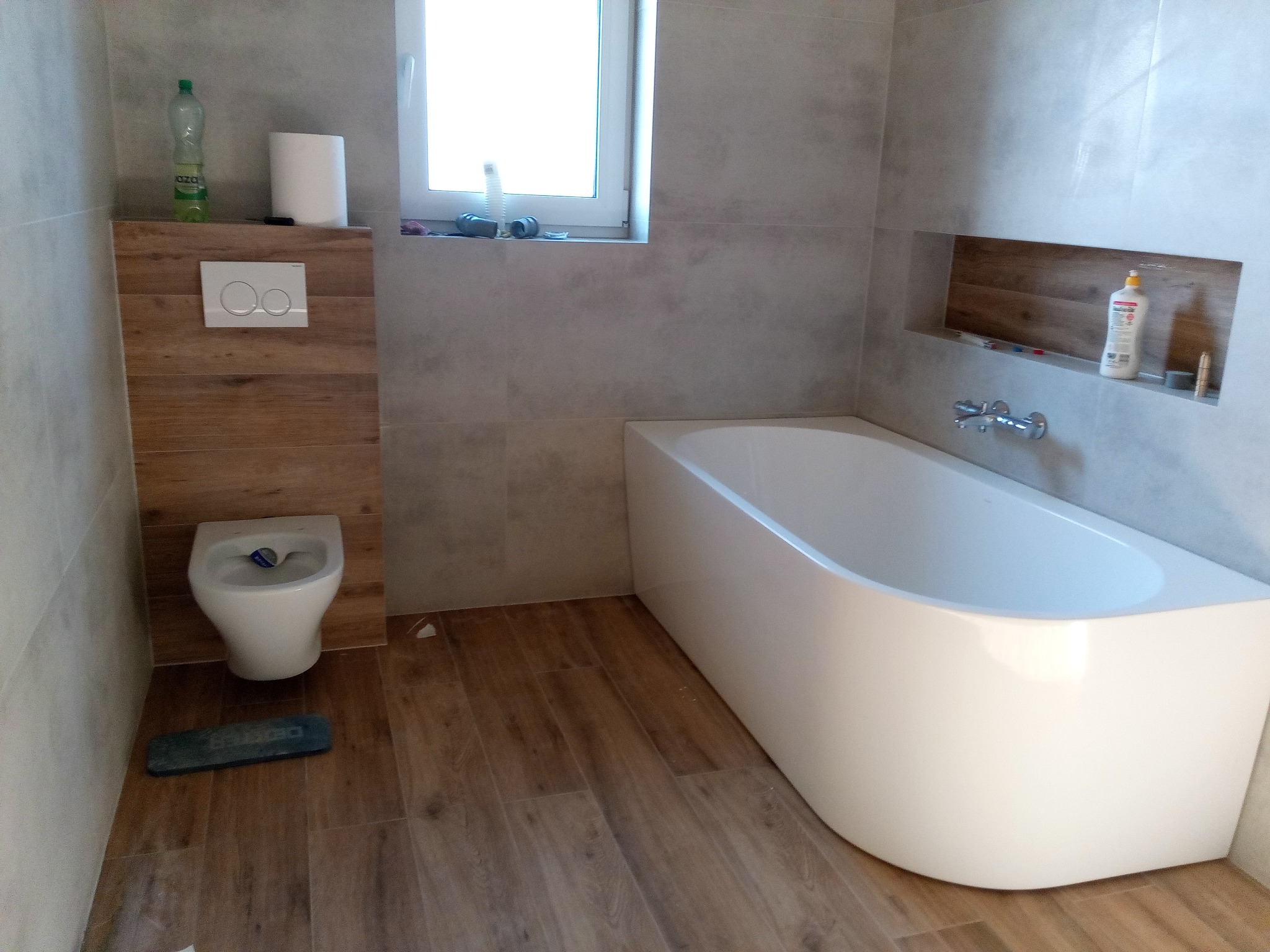 Wykończona łazienka z wanną, toaletą i drewnopodobnymi płytkami na podłodze i ścianie. Widoczne detale: armatura, okno z akcesoriami, półka w ścianie.