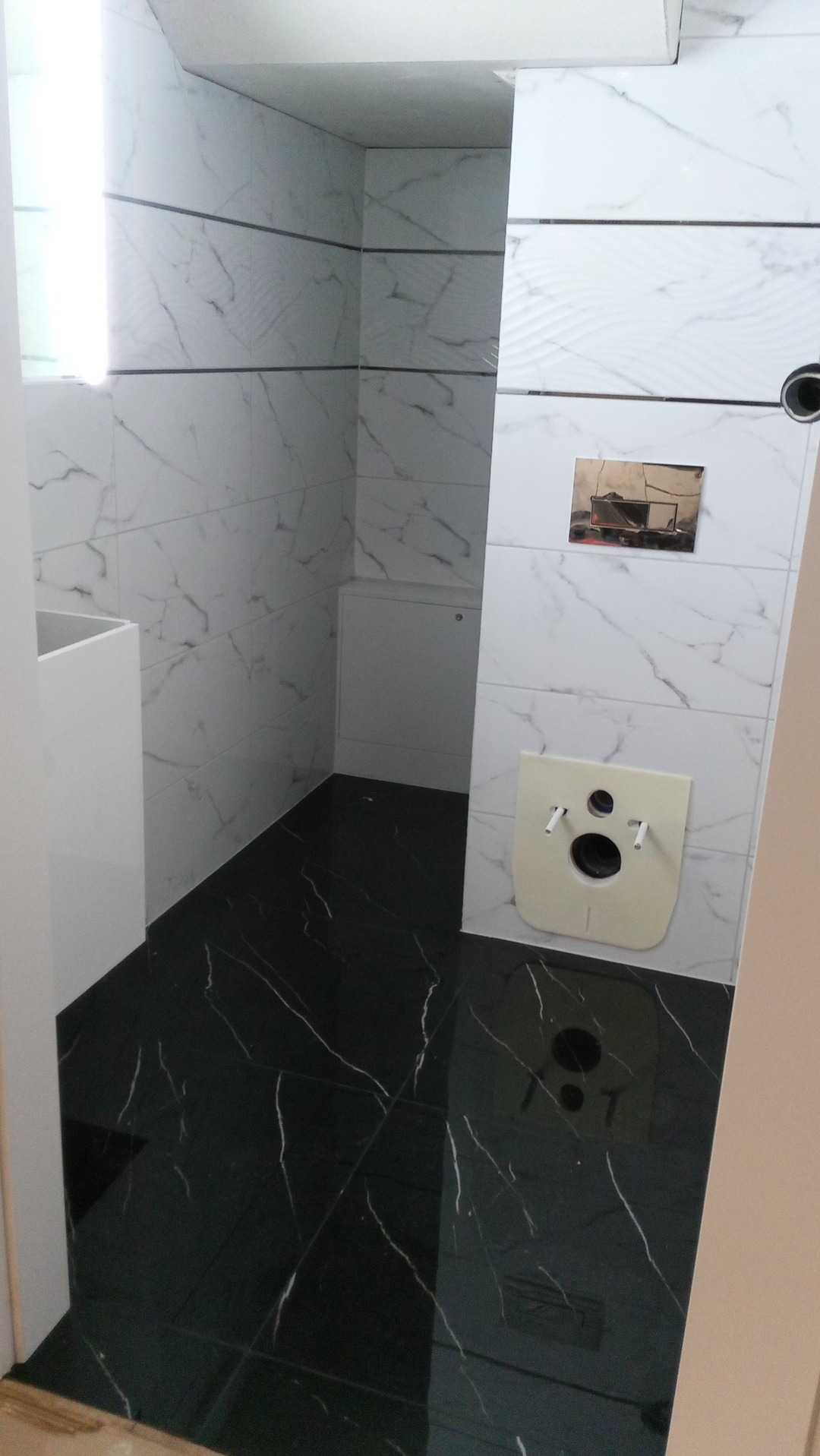 Nowoczesna łazienka z czarno-białymi płytkami imitującymi marmur. Widoczny stelaż podtynkowy WC i fragment umywalki. Minimalistyczny design.