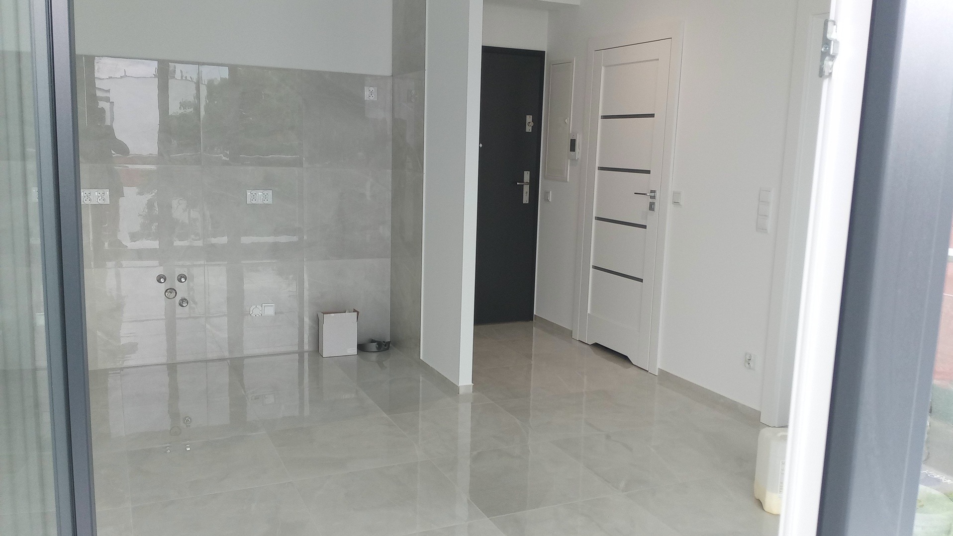 Wnętrze po remoncie z szarymi płytkami na ścianie i podłodze, widoczne gniazdka elektryczne i wyprowadzenia wodne. Dwa rodzaje drzwi w tle, białe ściany.
