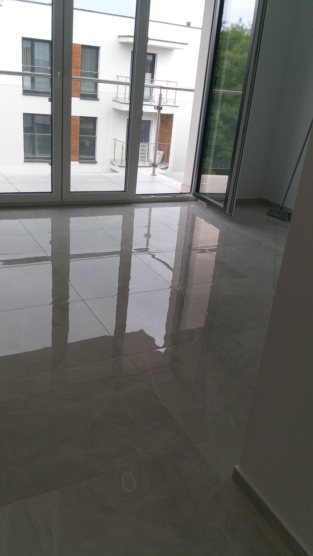 Błyszcząca, szara podłoga z płytek odbija światło z dużych okien balkonowych w nowoczesnym wnętrzu. Widoczny fragment ściany i sprzęt do sprzątania.