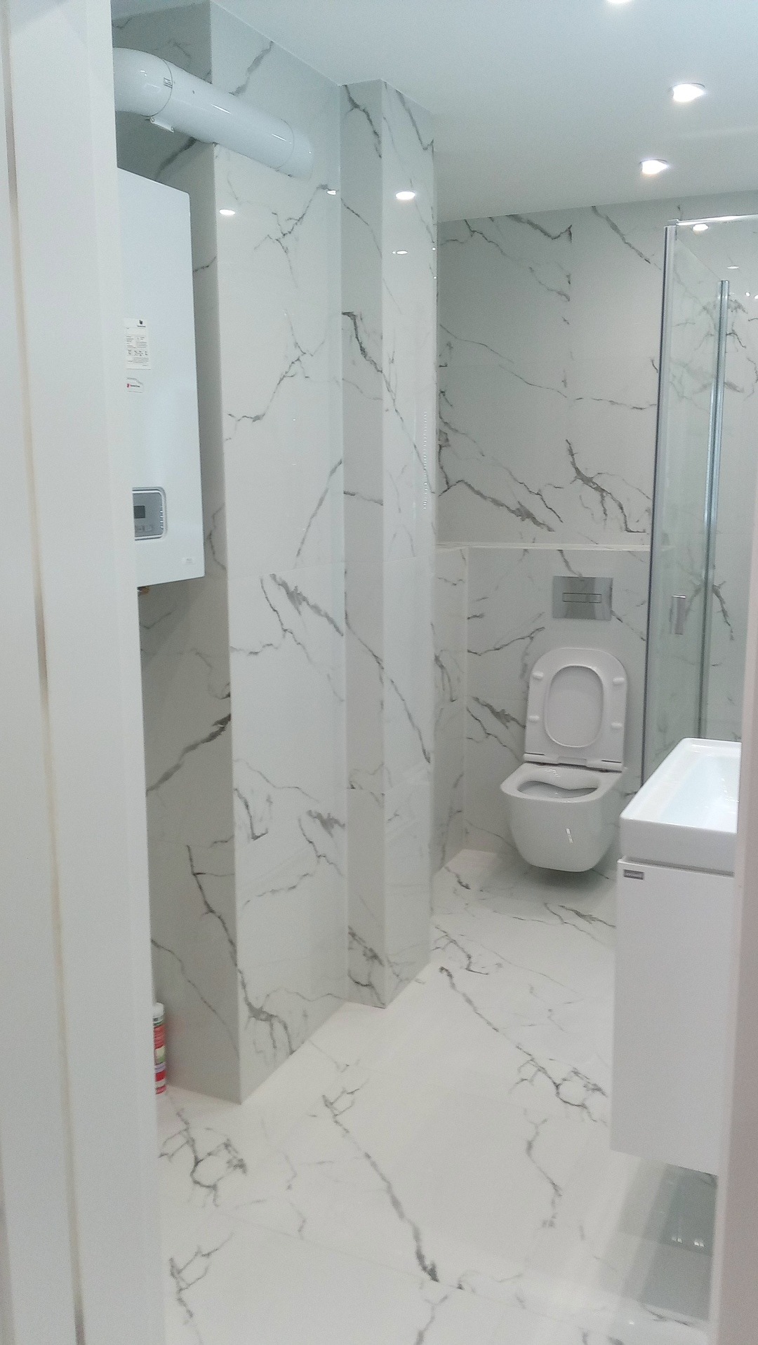 Wykończona łazienka w bieli z marmurowym wzorem na płytkach, widoczna kabina prysznicowa, toaleta i umywalka. Na ścianie piec gazowy. Całość w nowoczesnym stylu.