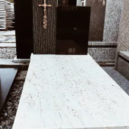 Czarny nagrobek z ozdobnym krzyżem i jasnym blatem, otoczony innymi nagrobkami, prezentowany w zakładzie kamieniarskim. Widoczne detale kamieni.