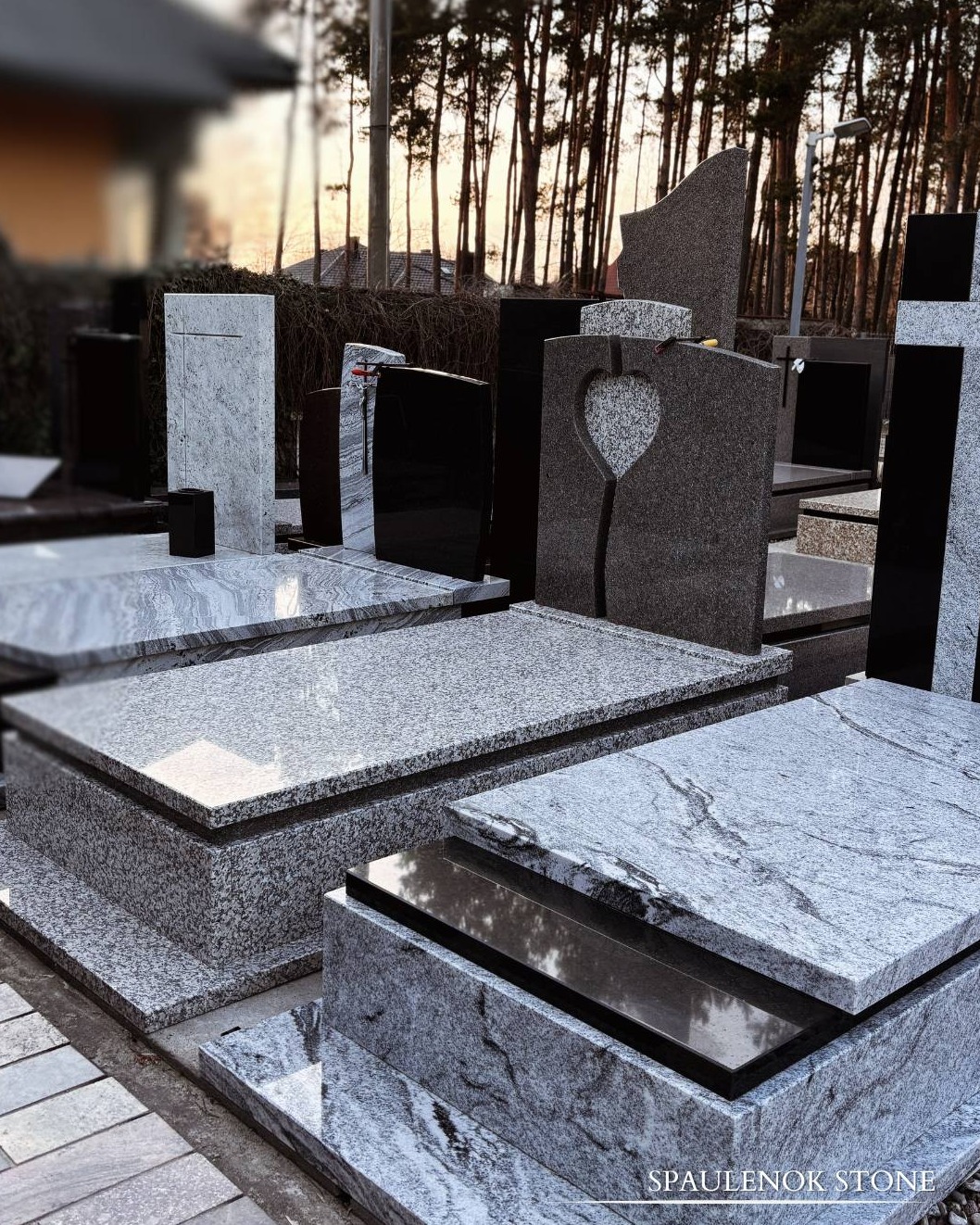 Różnorodne nagrobki z kamienia naturalnego i granitu, w odcieniach szarości i czerni, ustawione na ekspozycji zewnętrznej, z drzewami w tle.
