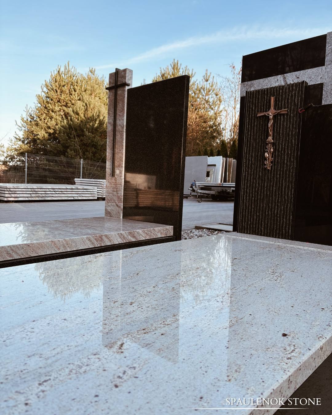 Wystawa nagrobków z krzyżami i ornamentami w Ostrowi Mazowieckiej. Różne wzory kamienia, widoczne odbicia. Ekspozycja w plenerze, błękitne niebo.