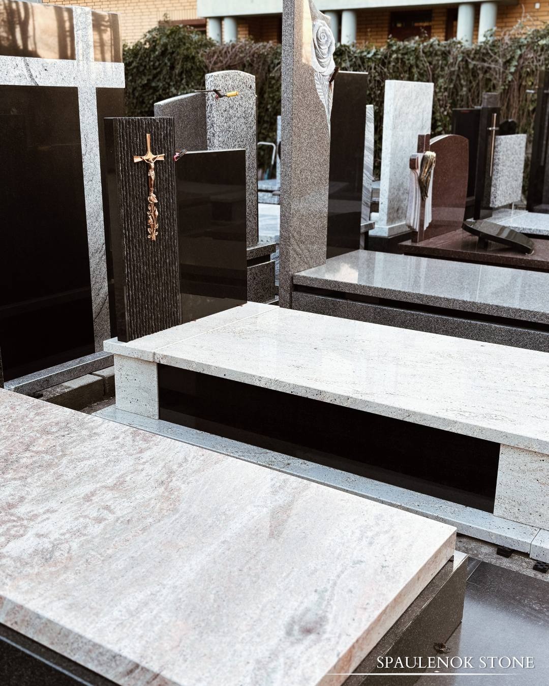 Wystawa nagrobków z granitu i marmuru o różnej kolorystyce i wzornictwie, z krzyżami i rzeźbami, na tle zieleni i budynku.