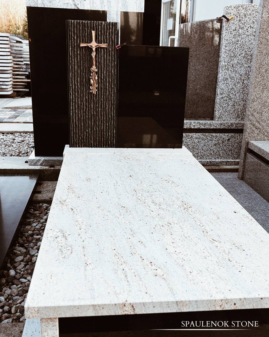 Czarny nagrobek z ozdobnym krzyżem i jasnym blatem, otoczony innymi nagrobkami, prezentowany w zakładzie kamieniarskim. Widoczne detale kamieni.