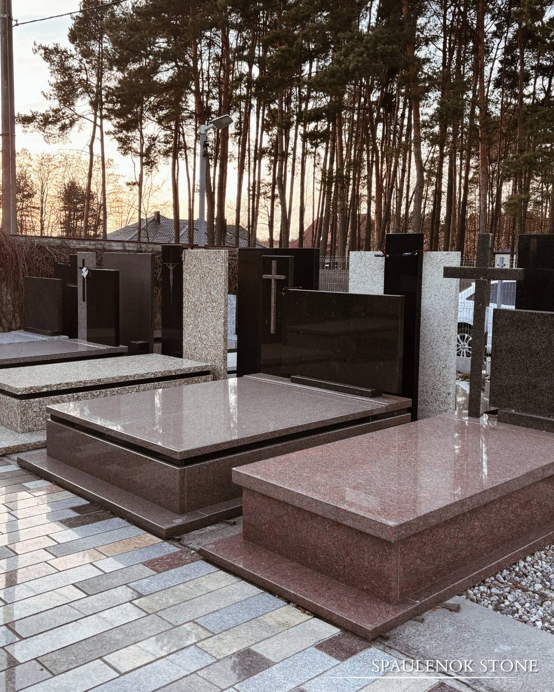 Granitowe nagrobki w odcieniach brązu i czerni, ustawione na kostce brukowej. W tle wysokie drzewa iglaste i fragment budynku. Widoczne różne wzory i wykończenia.