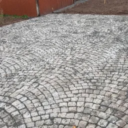 Ułożona z kostki granitowej nawierzchnia w kształcie wachlarza przed budynkiem z blachy falistej, widoczne świeżo przygotowane rabaty ziemi.