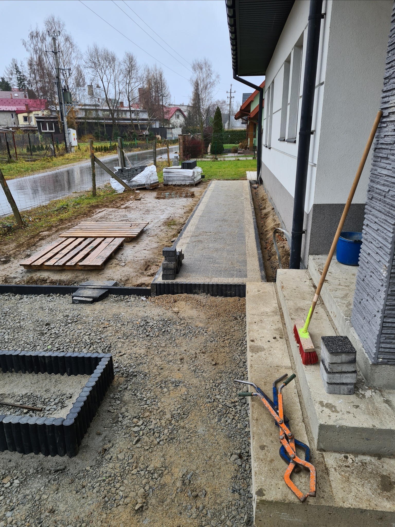 Etap prac: układanie szarej kostki na posesji domu jednorodzinnego, widoczne narzędzia brukarskie, obrzeża rabat i fragment ułożonego chodnika prowadzącego do wejścia.