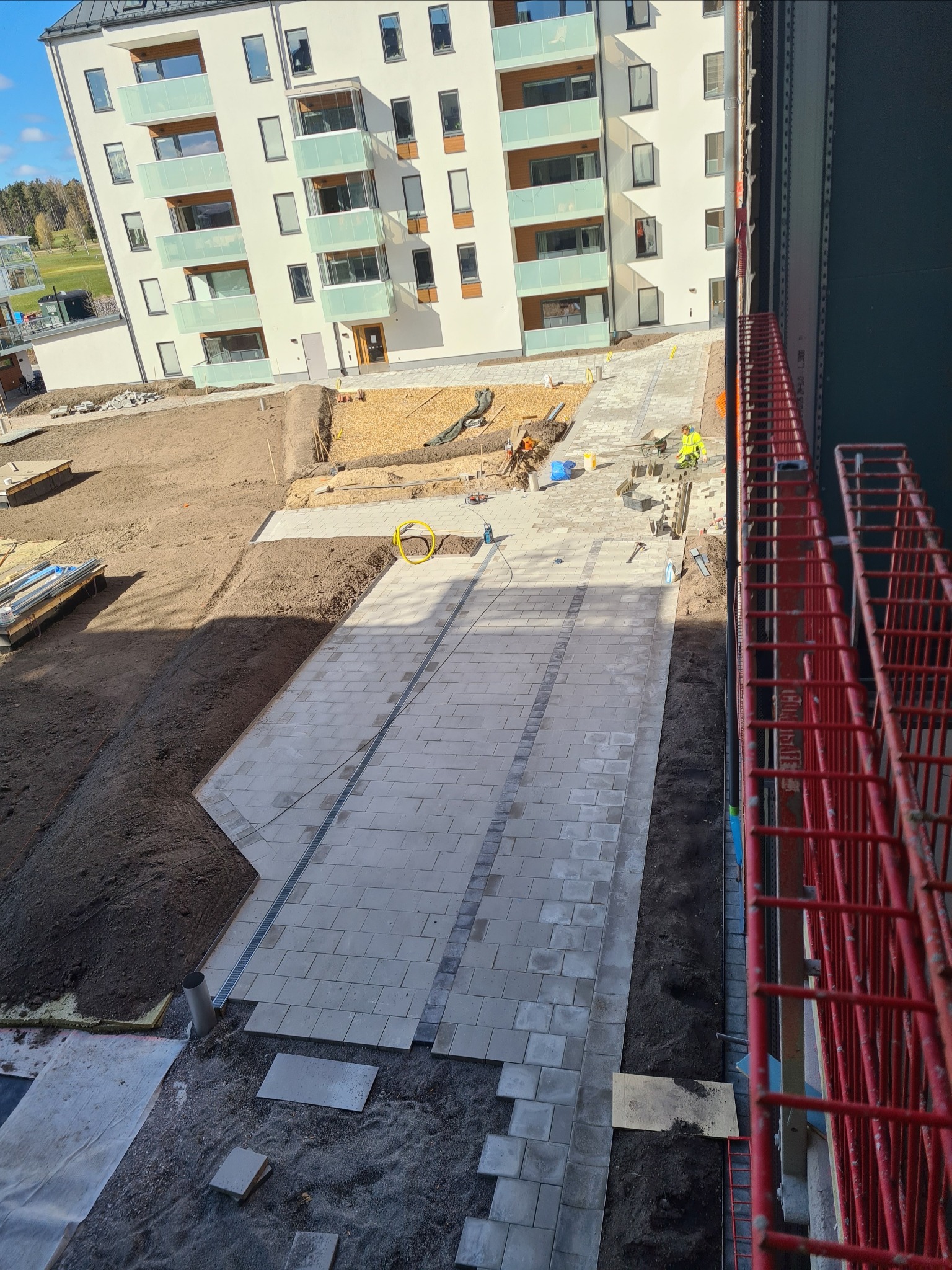 Widok z góry na plac budowy z częściowo ułożoną jasnoszarą kostką brukową, obrzeżami z ciemniejszej kostki, przygotowanym podłożem, pracownikami w odblaskowych kamizelkach i elementami rusztowania...