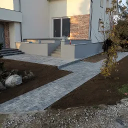 Nowoczesna posesja z szarą kostką brukową ułożoną w ścieżkę prowadzącą do domu z elementami cegły, świeżo obsadzona ziemią i roślinami.