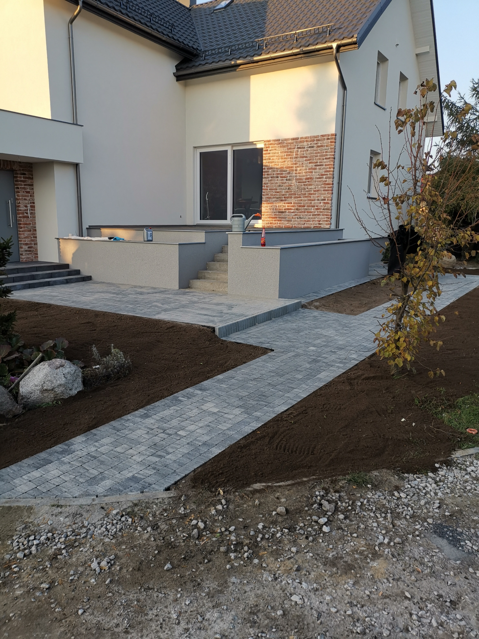 Nowoczesna posesja z szarą kostką brukową ułożoną w ścieżkę prowadzącą do domu z elementami cegły, świeżo obsadzona ziemią i roślinami.