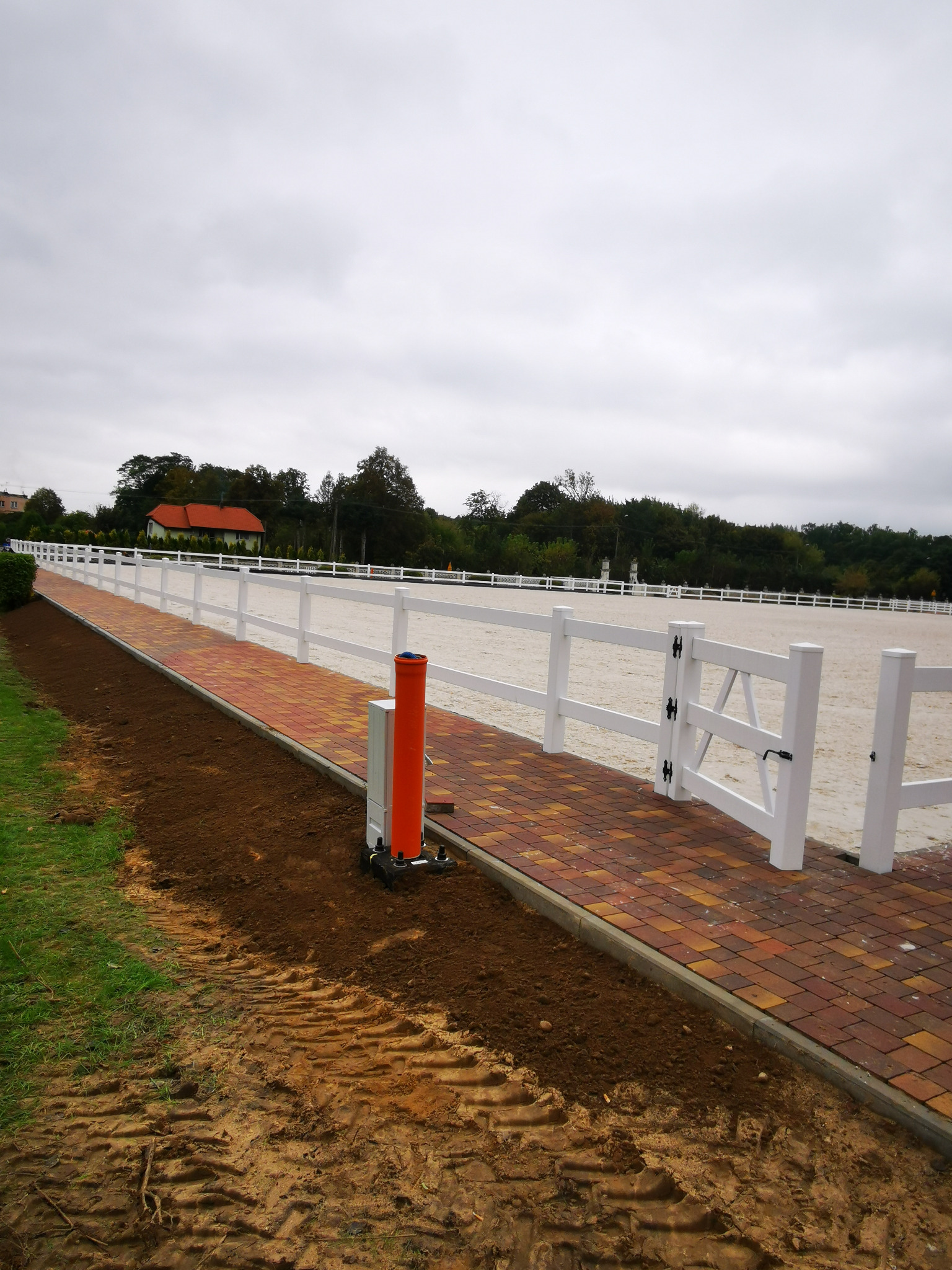 Wykończona nawierzchnia z kostki brukowej w kolorach jesieni, obok ogrodzenie terenu z białych paneli, świeżo obsiana trawa, pochmurne niebo.