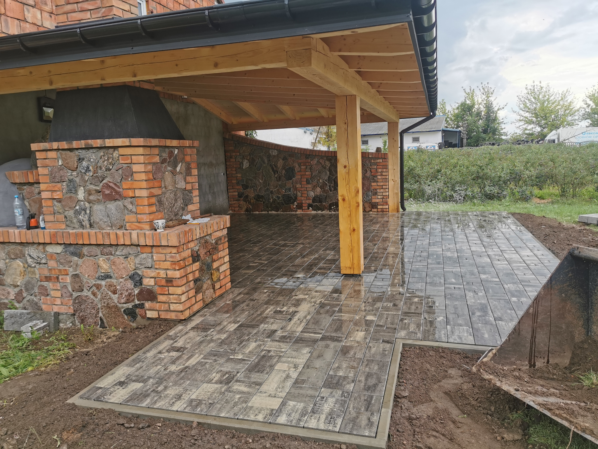 Świeżo ułożona, mokra kostka brukowa w Płocku, prowadząca do zadaszonej przestrzeni z ceglanym grillem. Widoczny fragment łyżki koparki i ziemia wokół nowej nawierzchni.