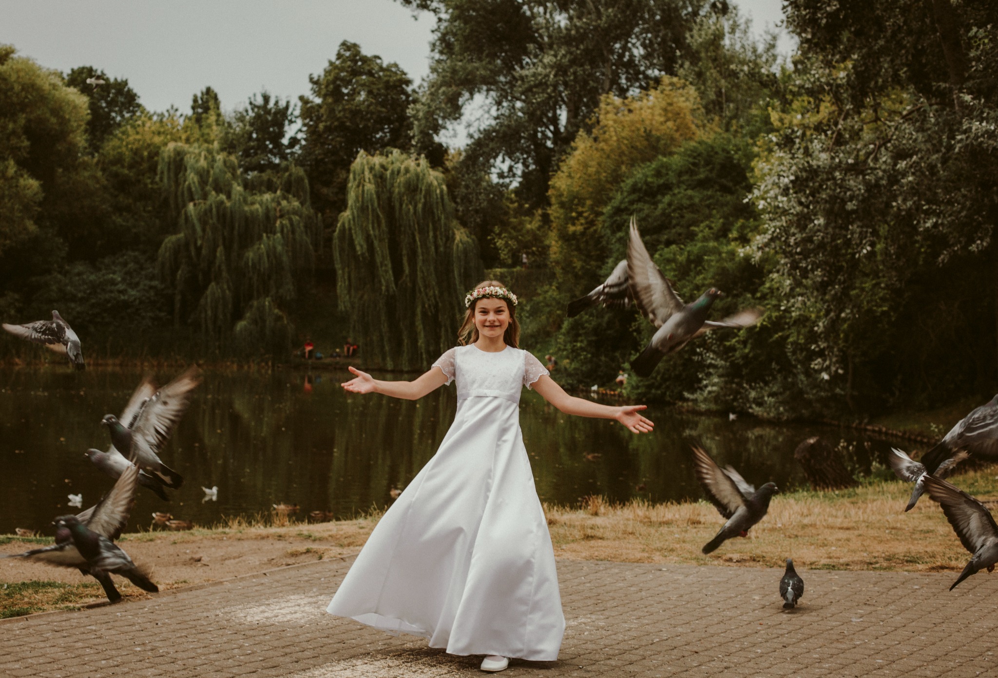 Dziewczynka w białej sukience i wianku na głowie, rozkłada ręce wśród stada gołębi na tle parku z jeziorem.