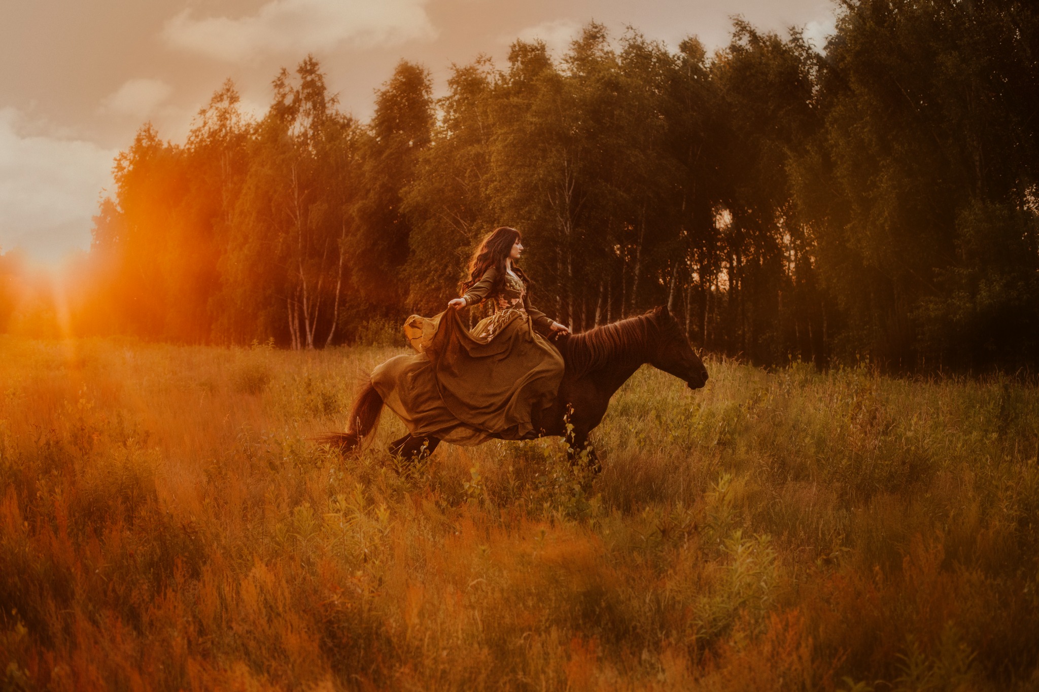 Kobieta w długiej sukni galopuje na brązowym koniu przez zarośnięte pole o zachodzie słońca; w tle widoczny las.