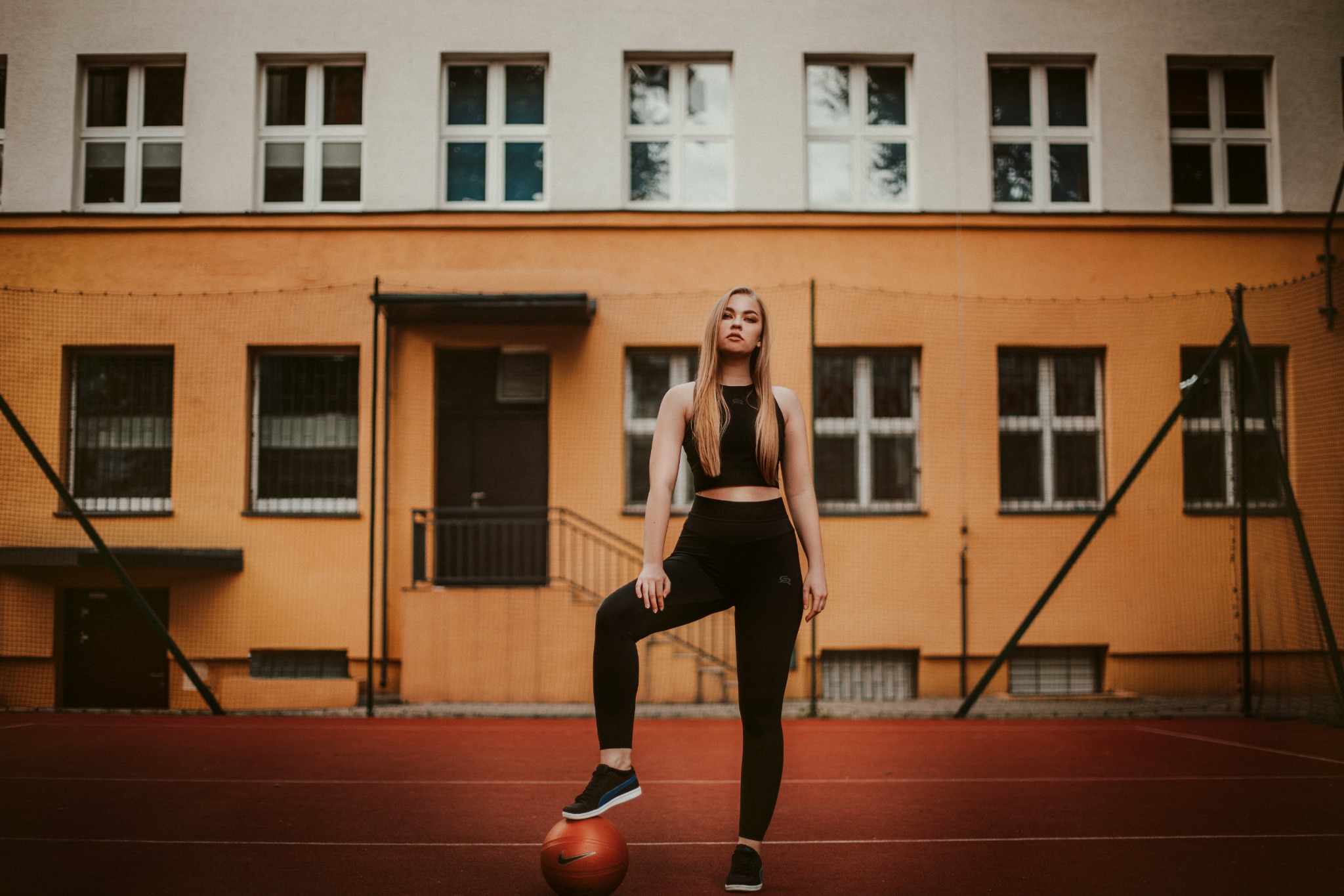 Młoda kobieta w sportowym stroju pozuje z piłką do koszykówki na tle budynku szkolnego z siatką ochronną.